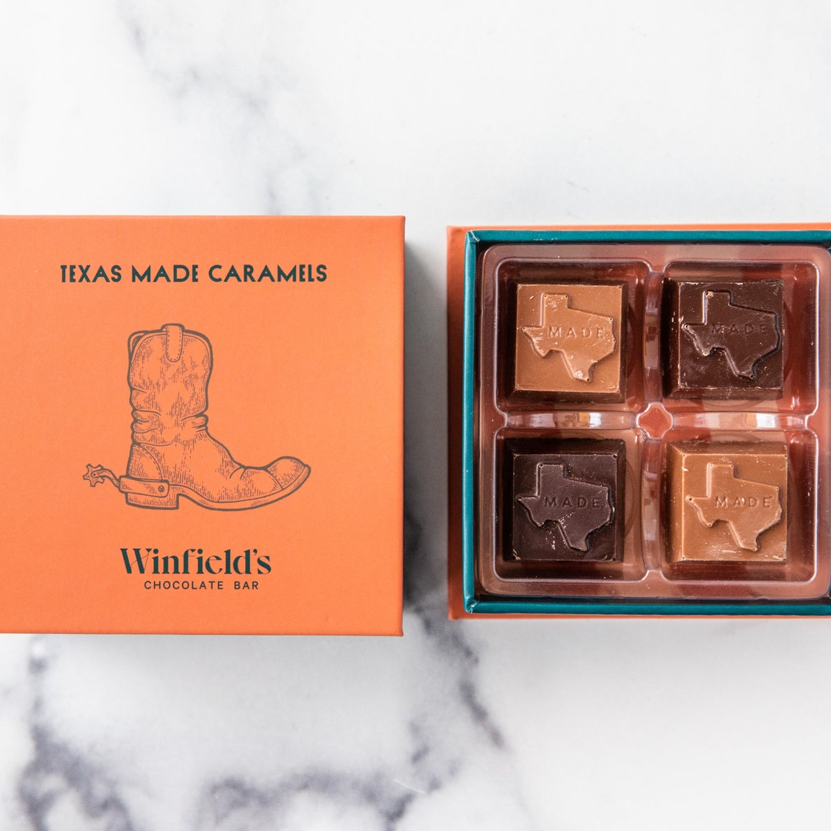 Winfield's Texas-Made Caramels 4-piece box in orange with a cowboy-boot crest, beside an open tray of four Texas-shaped chocolate caramels