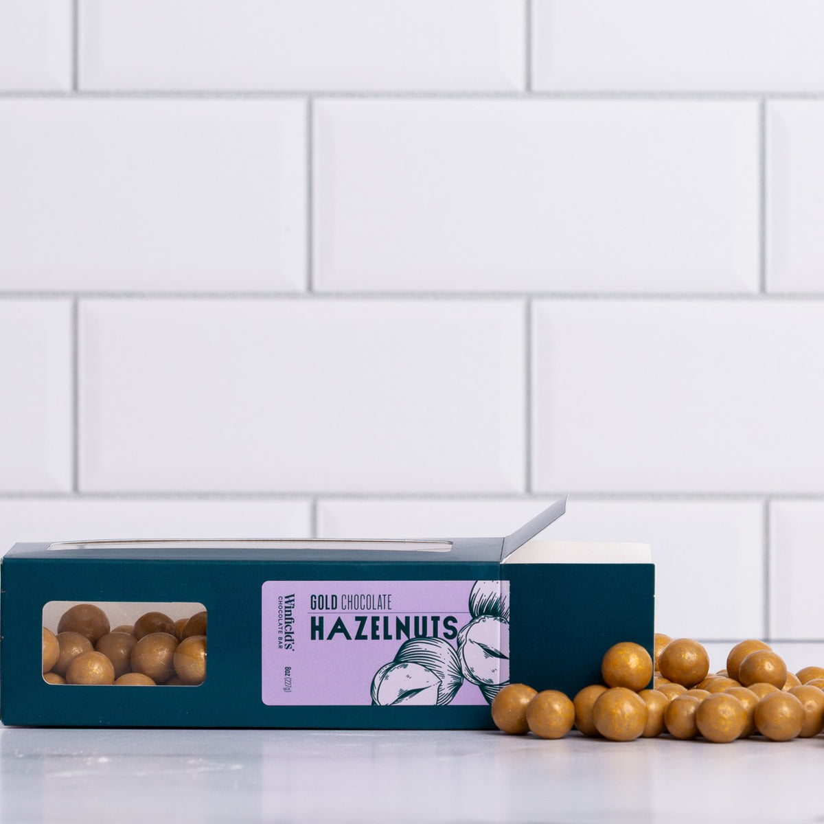 Winfield's Gold Chocolate Hazelnuts in a teal window box with loose gold-dusted hazelnuts alongside