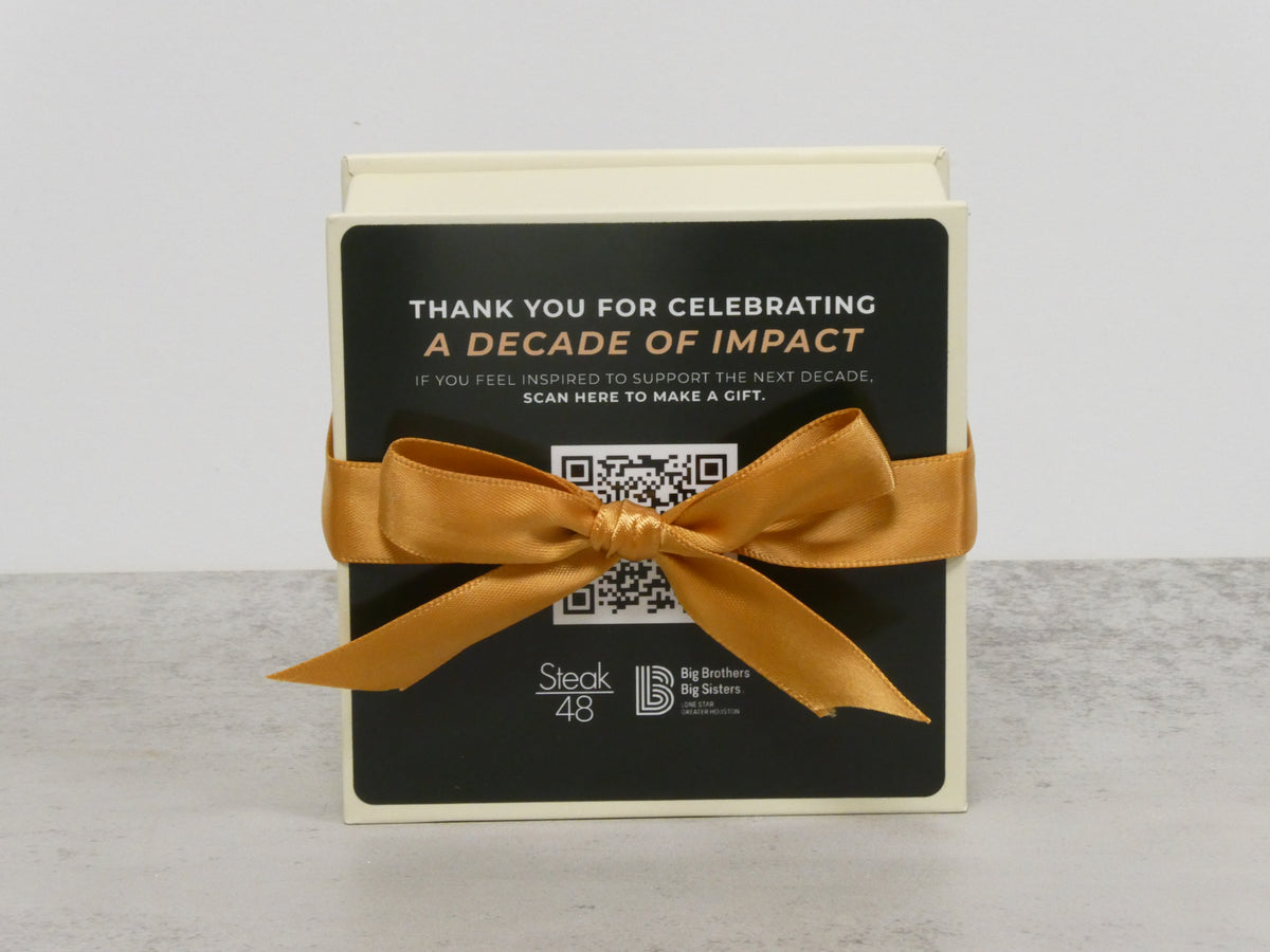A black Winfield's gift box tied with gold ribbon, its card reading Thank you for celebrating a decade of impact beside a QR code
