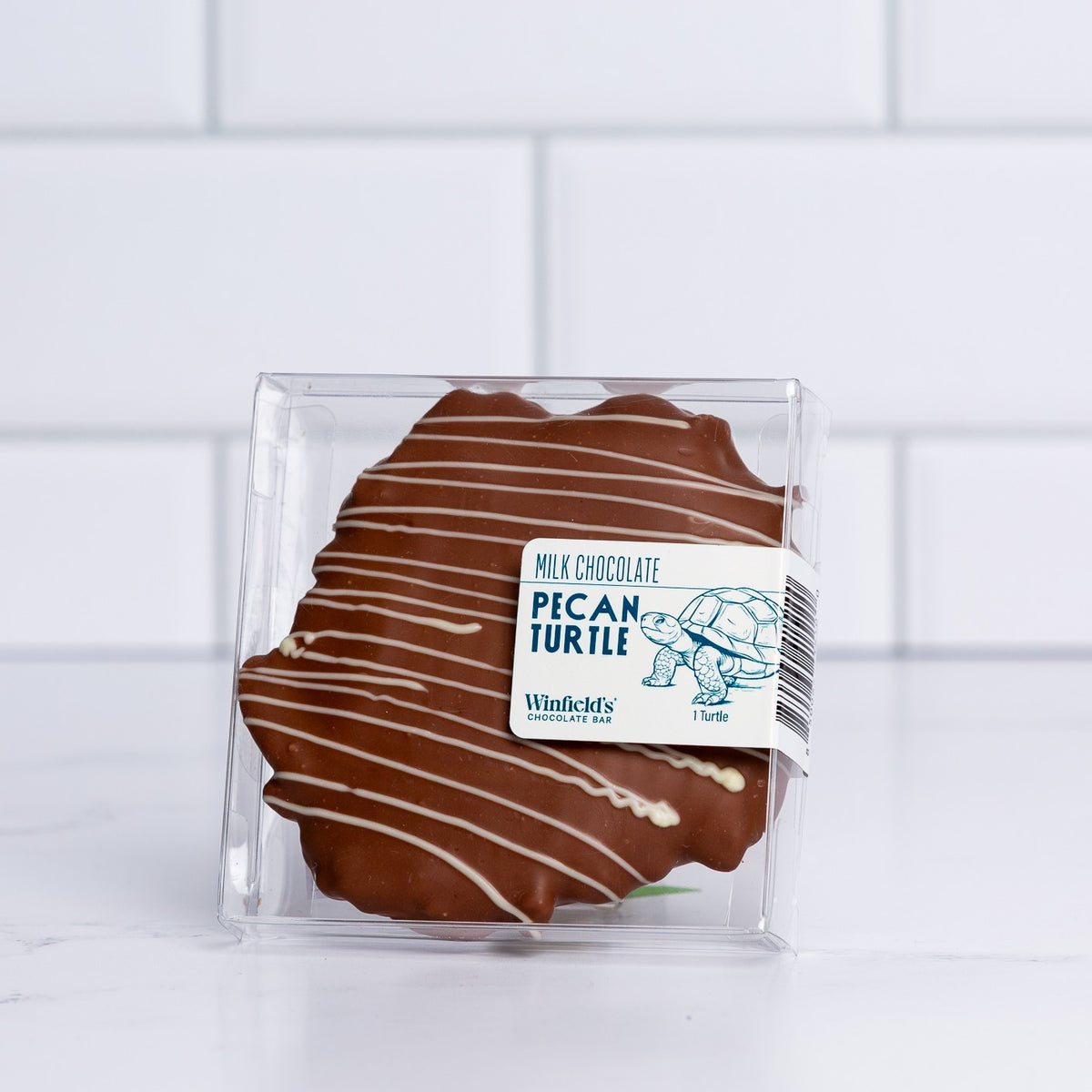 A single Winfield's milk chocolate pecan turtle with white chocolate drizzle in a clear gift box