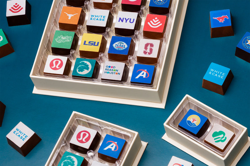 Open boxes of Winfield's chocolate squares, each square printed with a university or company logo, on a teal background