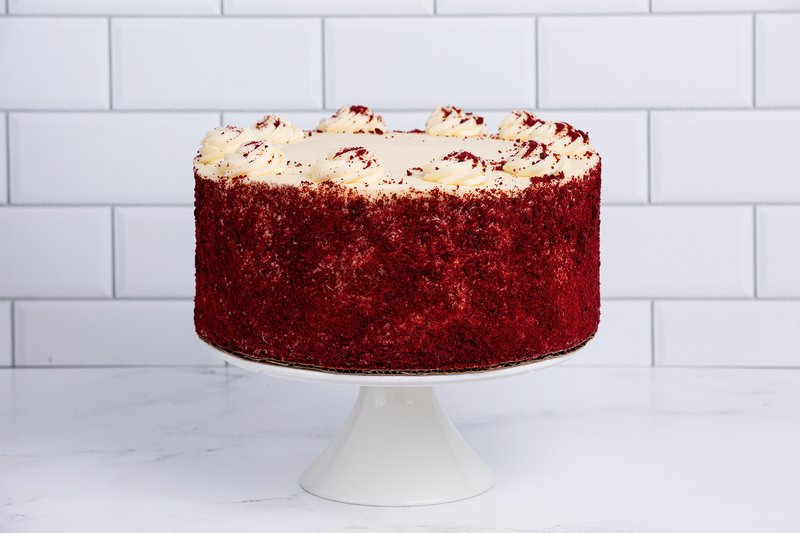 A whole Winfield's red velvet cake on a white cake stand, finished with cream cheese frosting and red crumb
