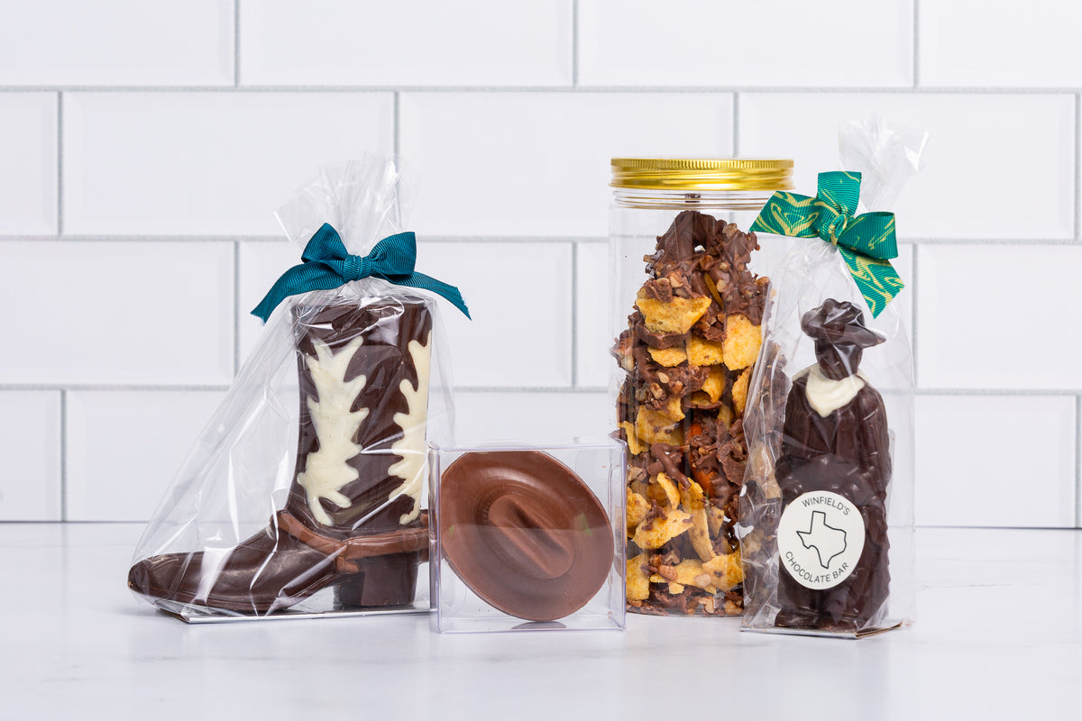 A molded chocolate cowboy boot and a bag of Texas-shaped chocolates tied with teal and gold ribbon against white subway tile