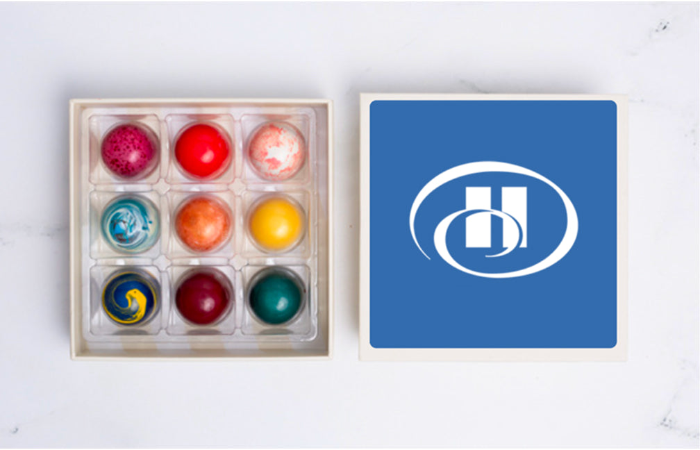 A nine-piece Winfield's truffle box beside a blue tile printed with a client logo, showing how custom branding is paired with a gift
